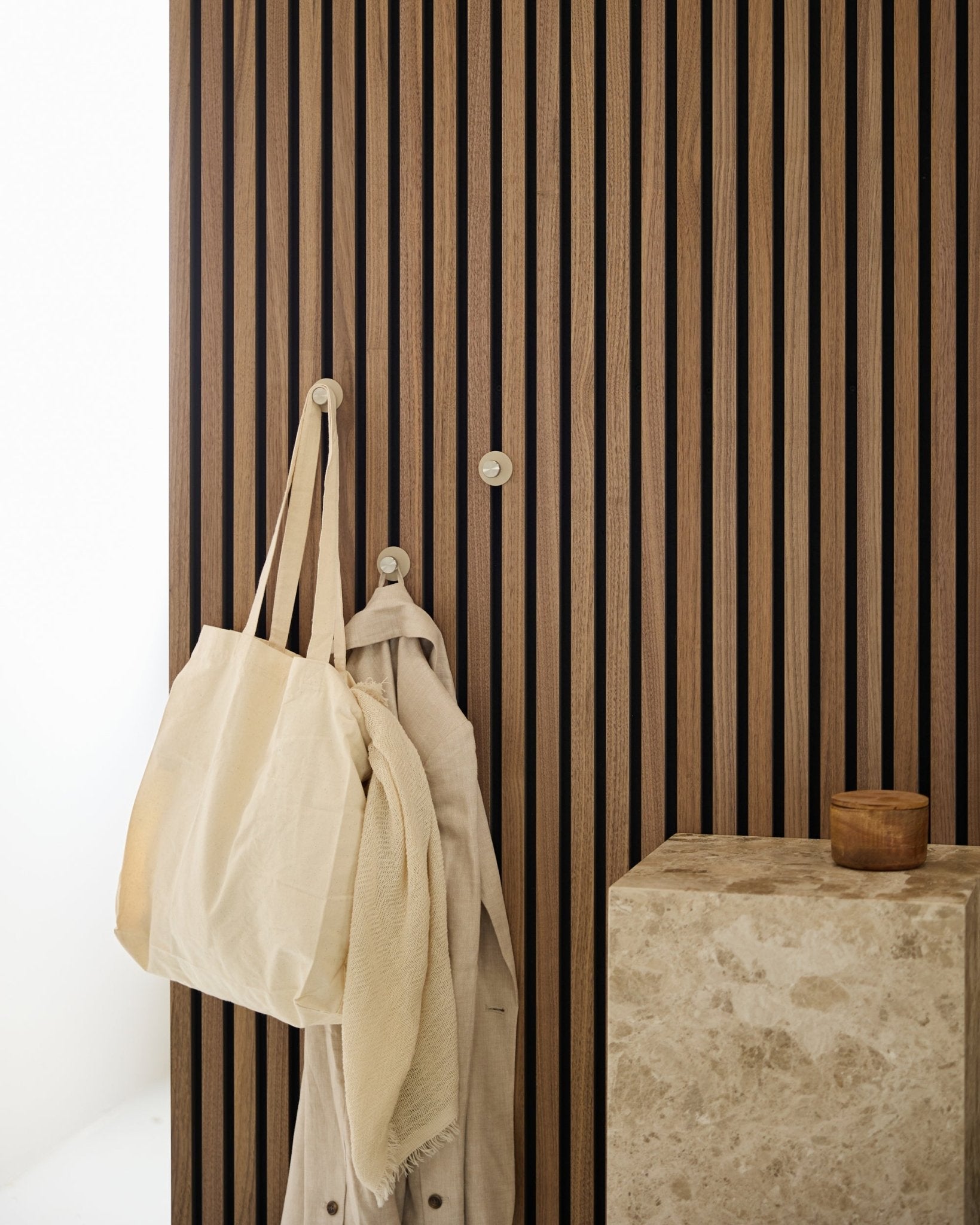 En beige tote bag og et lyst stoff henger fra en WoodUpp Norr knagg (Beige/rustfritt Steel) montert på en vertical Akupanel Akustikkpanel ved siden av en steinoverflate.