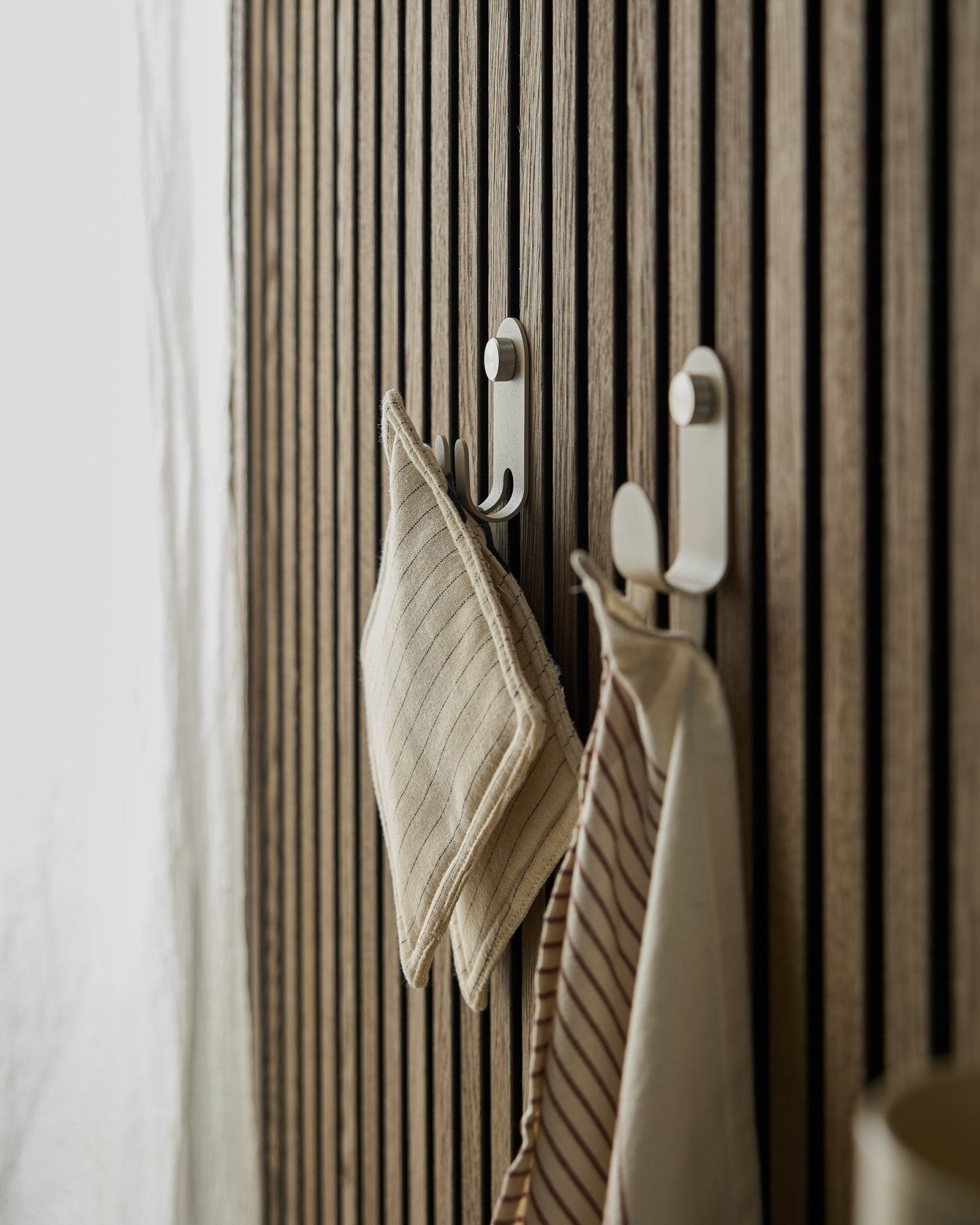 To stripete kjøkkenhåndklær henger på Norr Double Hanger fra WoodUpp, montert på et beige nordisk spilepanel Akustikkpanel, opplyst av mykt naturlig lys.