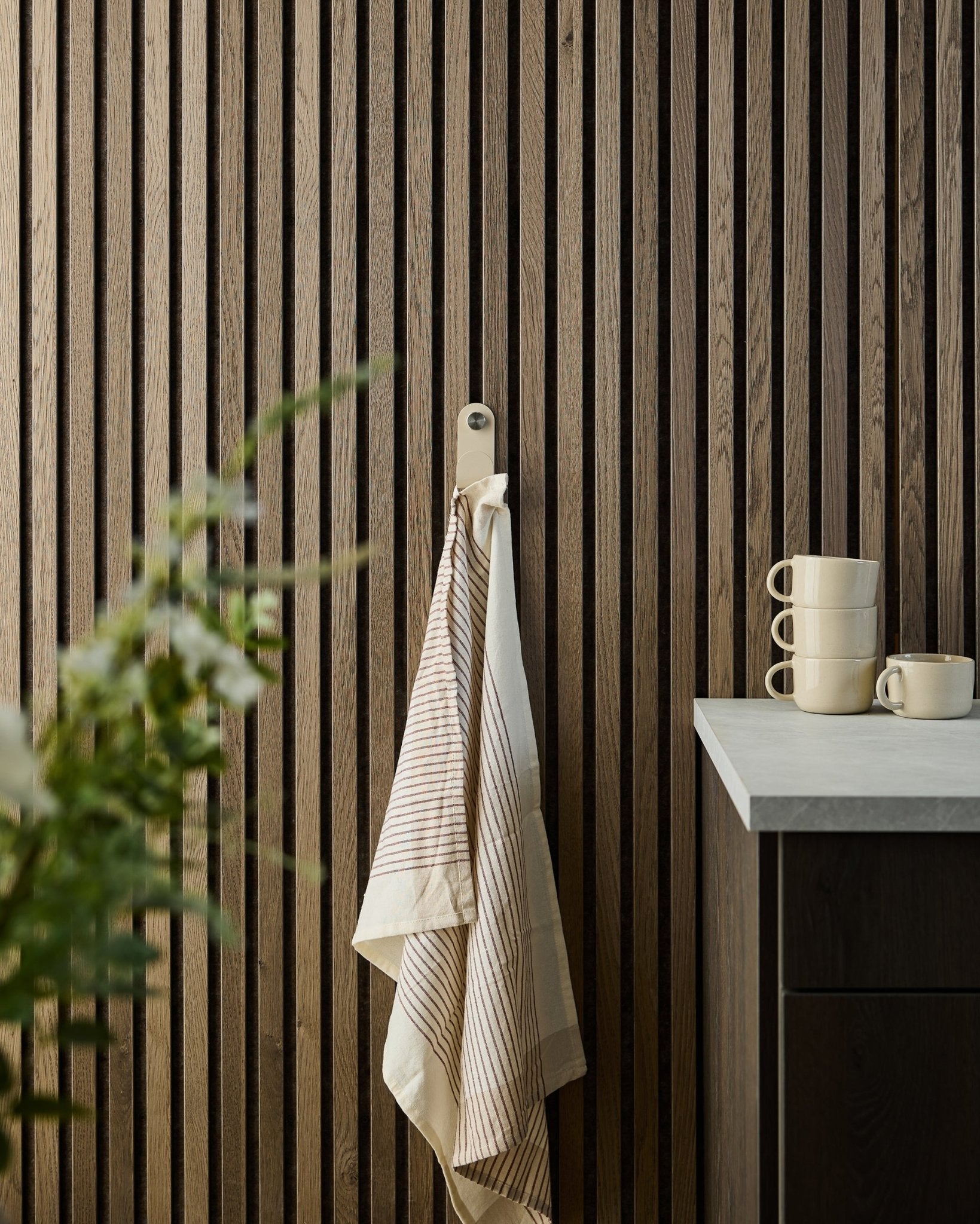 Et stripete håndkle henger fra en beige WoodUpp Norr Single Hanger montert på et veggpanel med trespiler Akustikkpanel; stablede krus og en plante står på benken under.