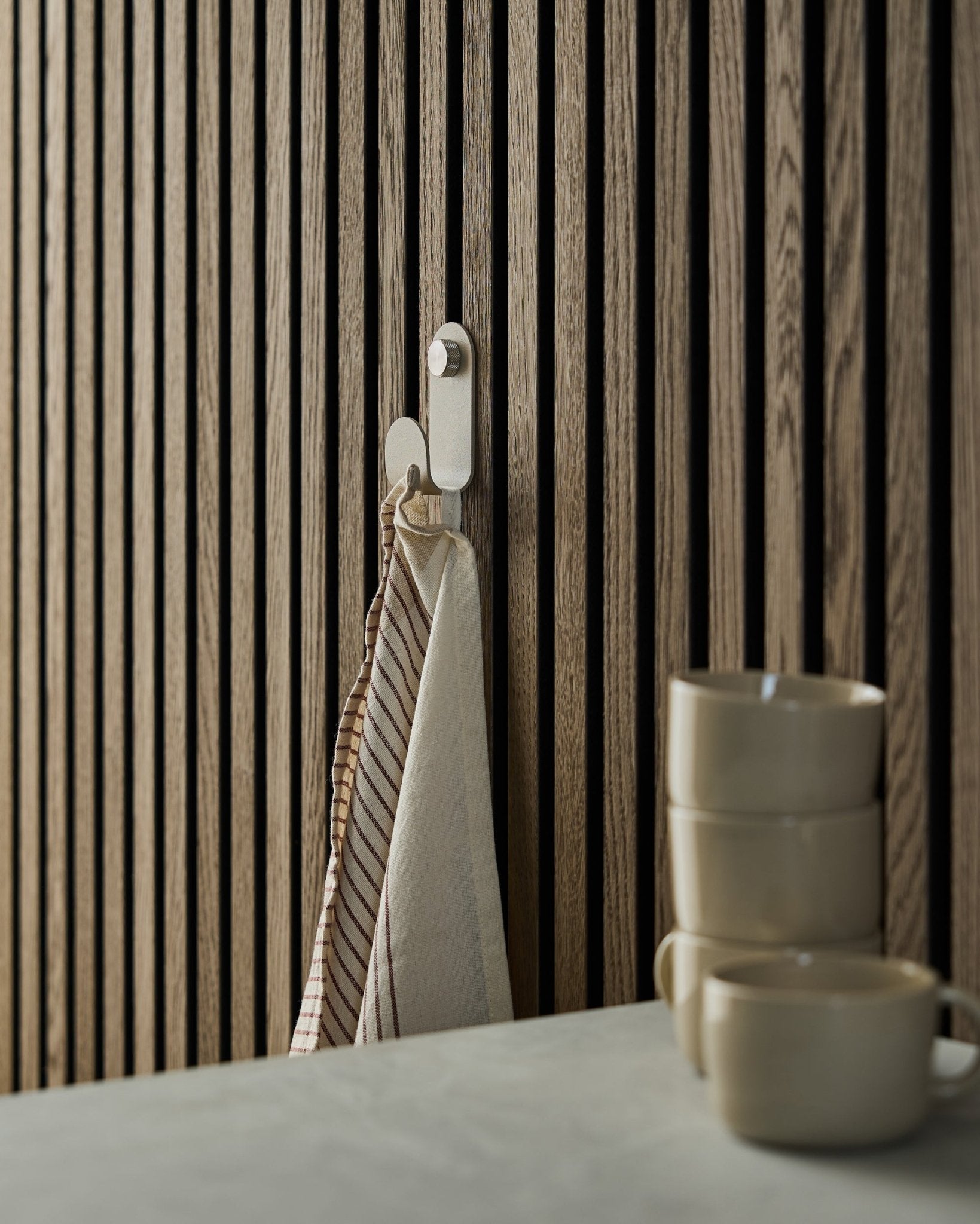 Et stripete håndkle henger fra en WoodUpp Norr Single Hanger i beige, montert på et akustisk spilepanel i nordisk stil; tre beige krus er stablet foran.