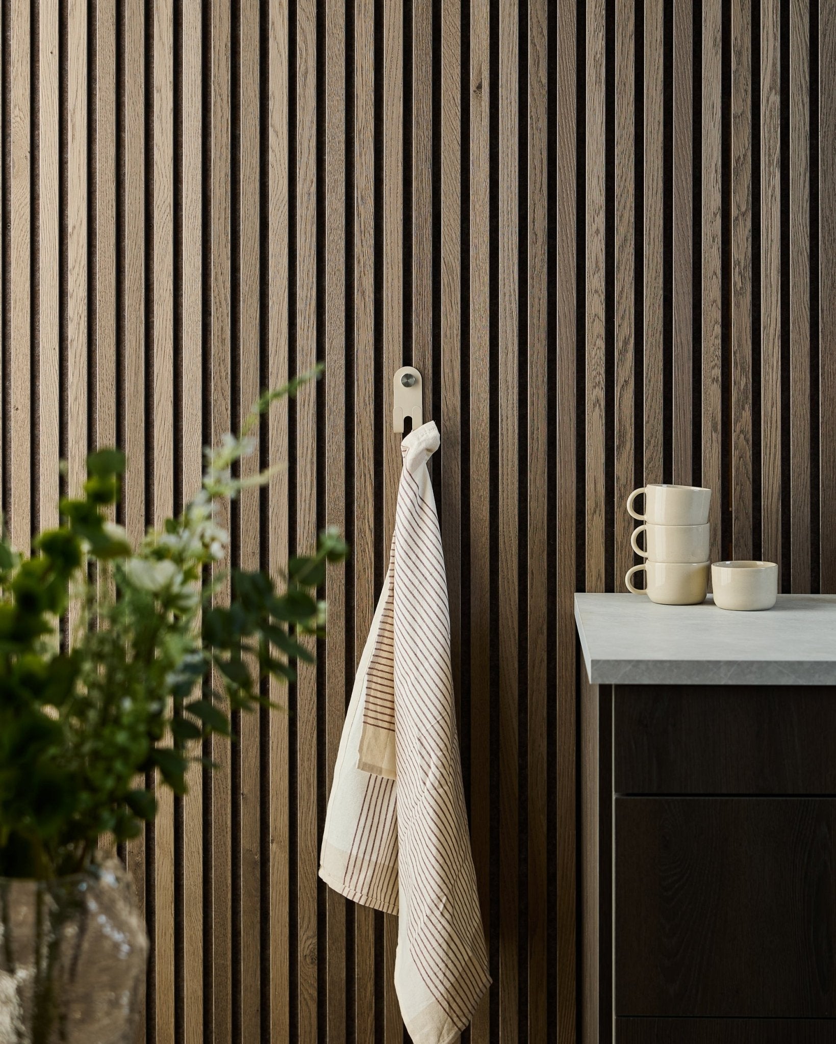 Et stripete håndkle henger på WoodUpp Norr Double Hanger i beige, montert på et nordisk akustisk spilepanel; kopper, en bolle og en grønn plante står under.