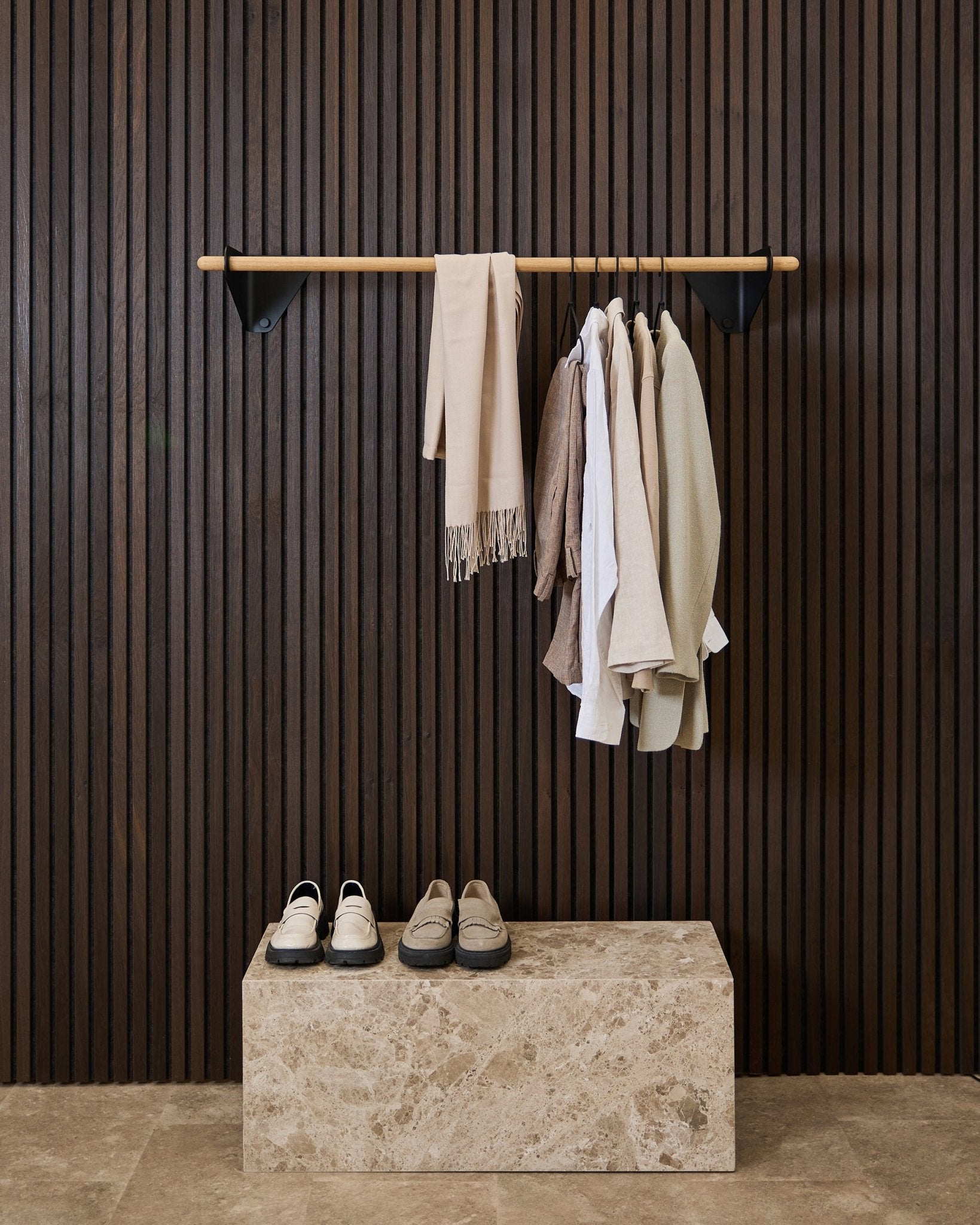 Klær i nøytrale toner og et skjerf henger på en black 115,9 cm Linn | Coat Rail fra WoodUpp over en benk i marmor, mot et mørkt Akupanel akustisk spilepanel.