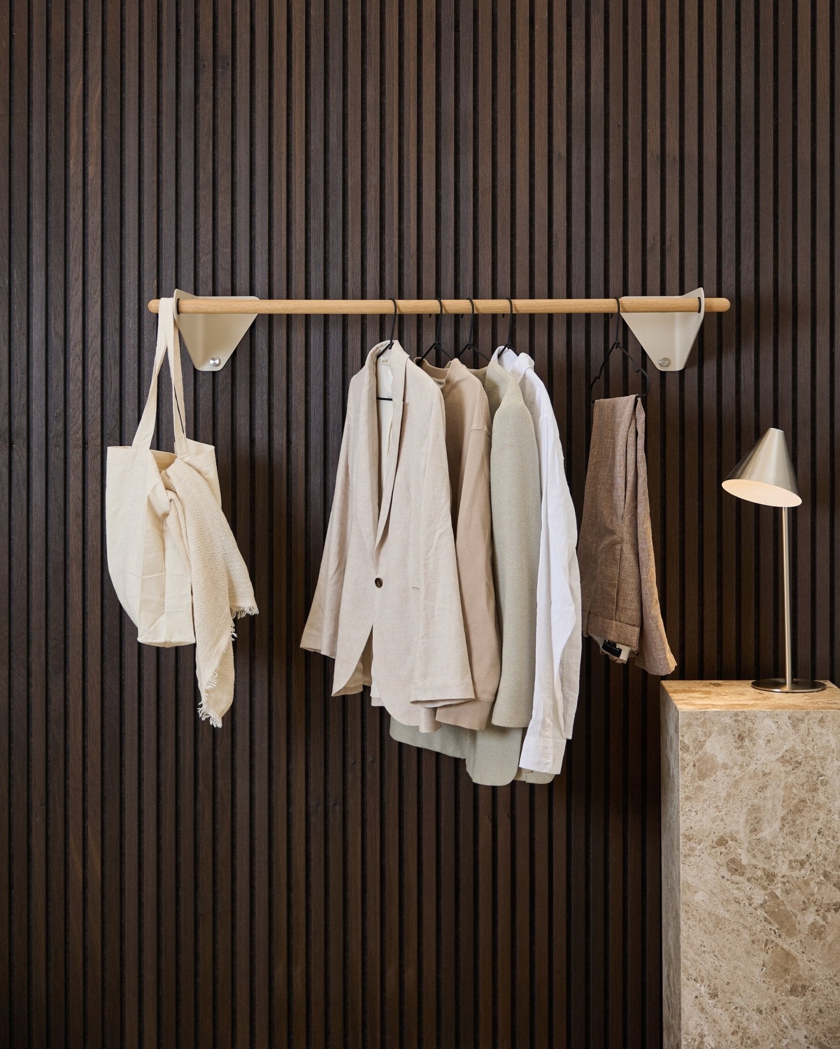 En beige WoodUpp Linn klesstang (115,9 cm) med jakker, en hvit skjorte og en kremfarget totebag foran et mørkt tre Akupanel Akustikkpanel og en marmorlampе.