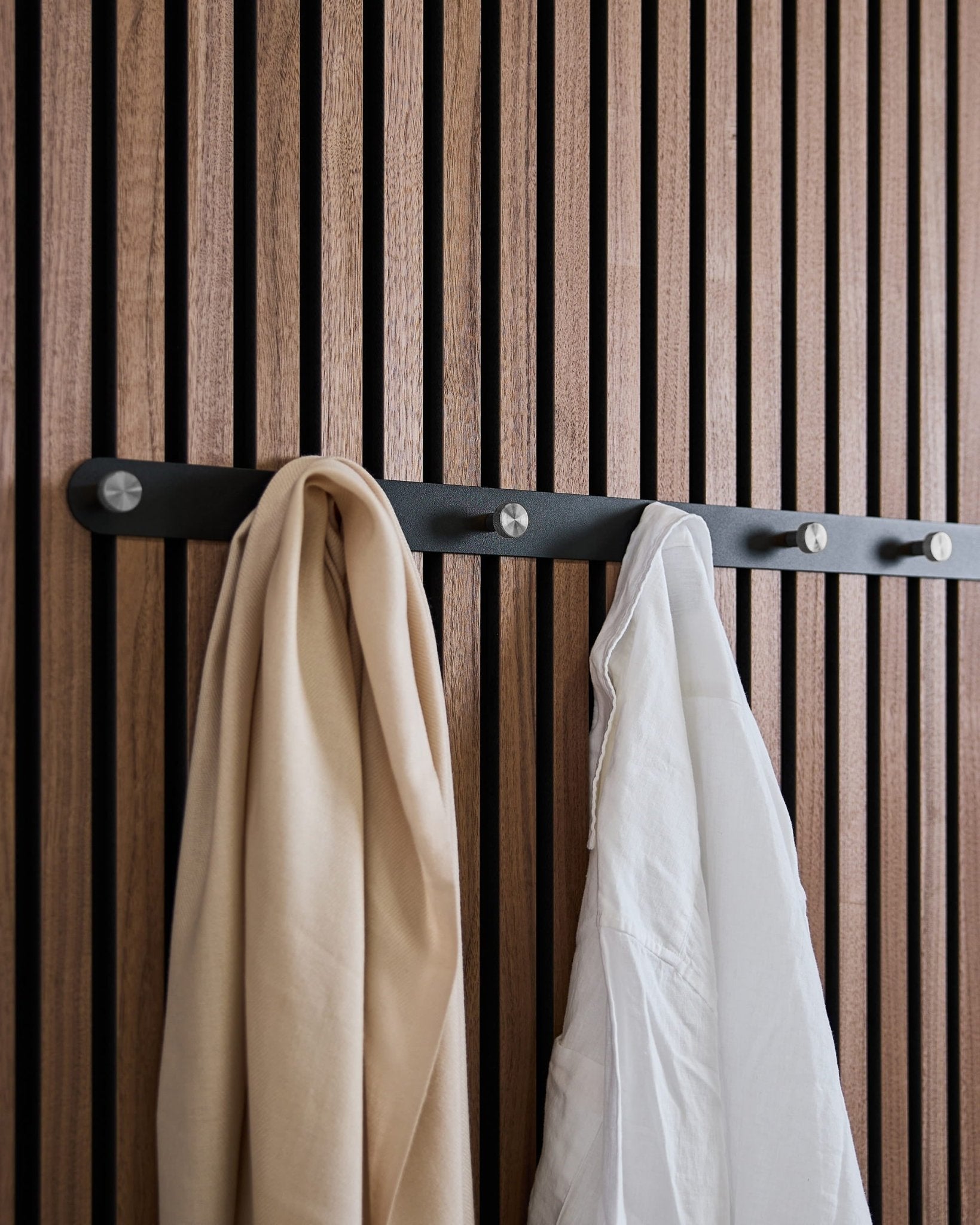 Et beige skjerf og en hvit skjorte henger på WoodUpps Norr X7 Black/Stainless Steel knaggrekke, montert på et nordisk akustisk spilepanel.