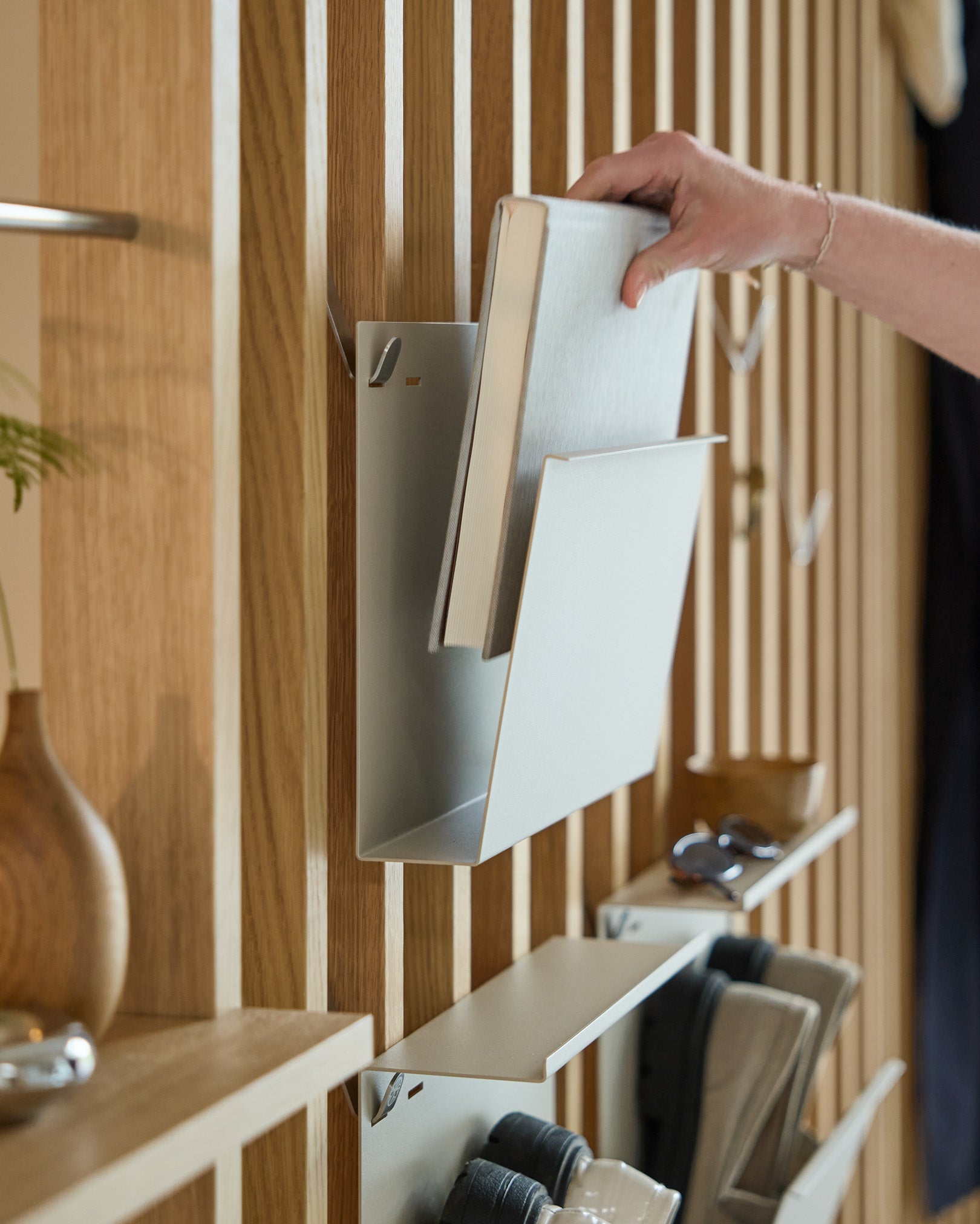En hånd plasserer Nell | Magazine Storage / Beige fra WoodUpp på en pulverlakkert hylle i steel, montert på en nordisk trebasert Akustikkpanel, med gjenstander i nærheten.