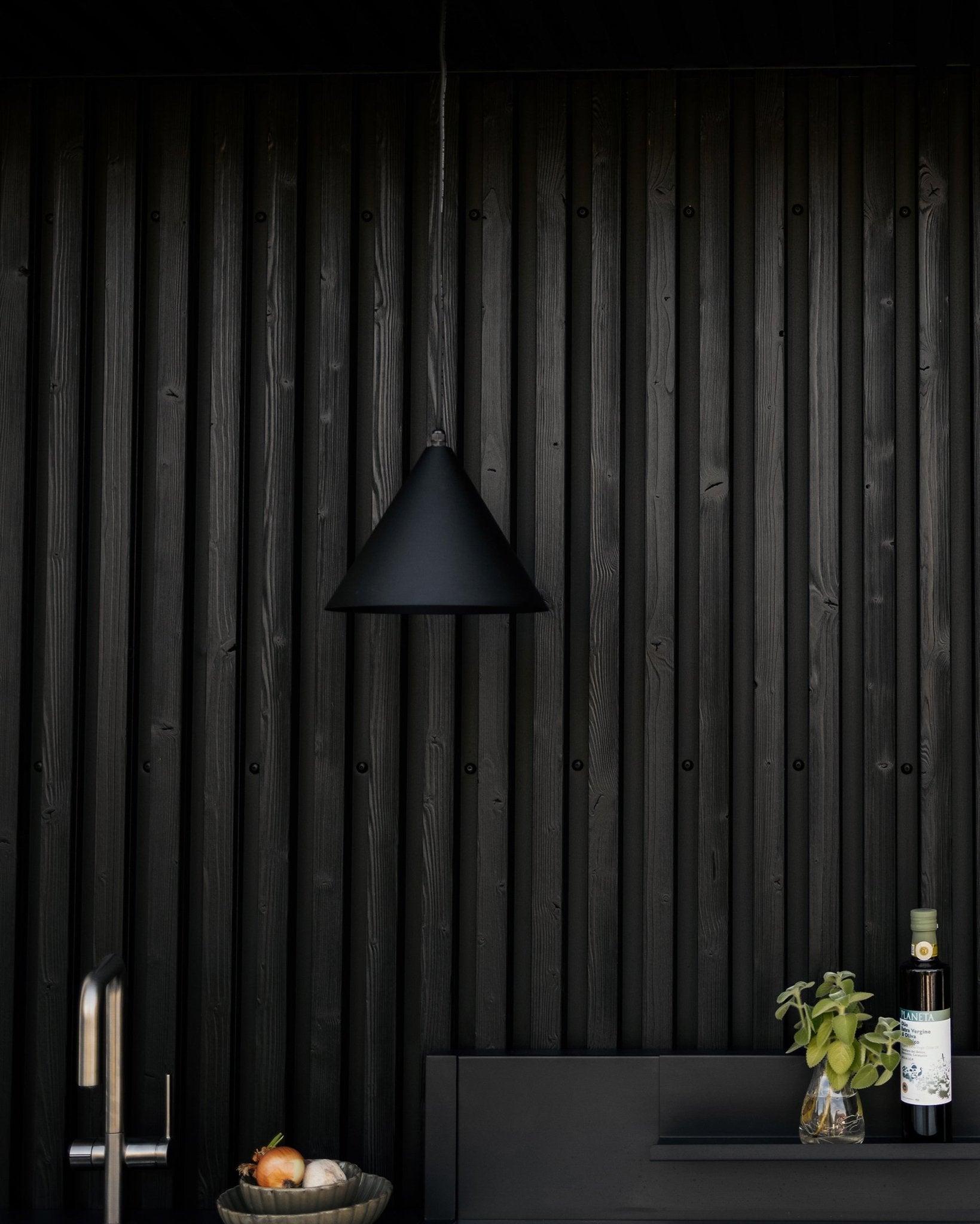 Moderne kjøkken med pendellampe i matt black, teksturert akustisk trevegg med black Akupanel, og benkeplate med grønnsaker, fersk basilikum og olivenolje.