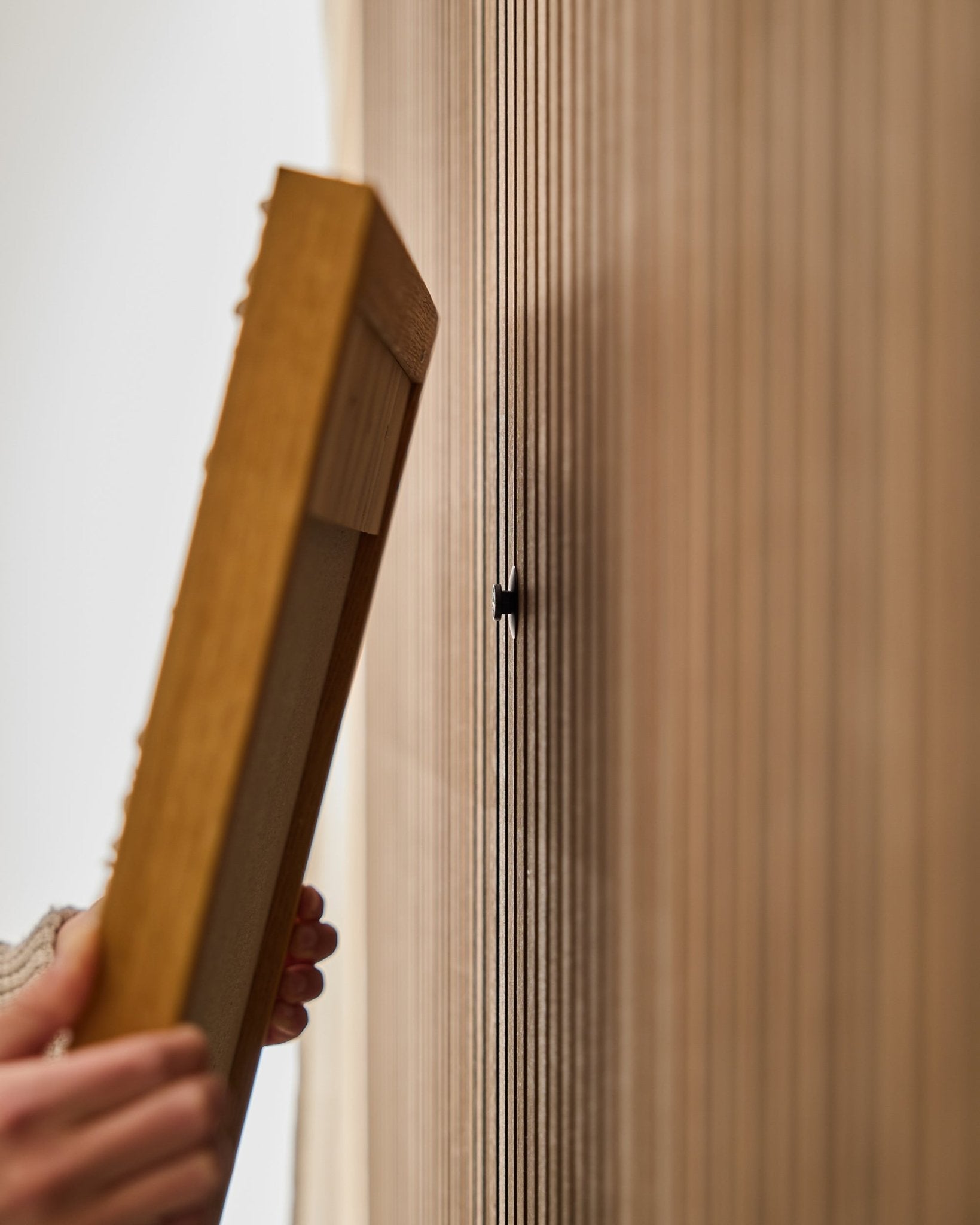 En person bruker WoodUpp Dupps | Original-settet for å montere en ramme til et Akupanel akustisk trepanel, med fokus på å feste en small black krok nær vertical spilene.