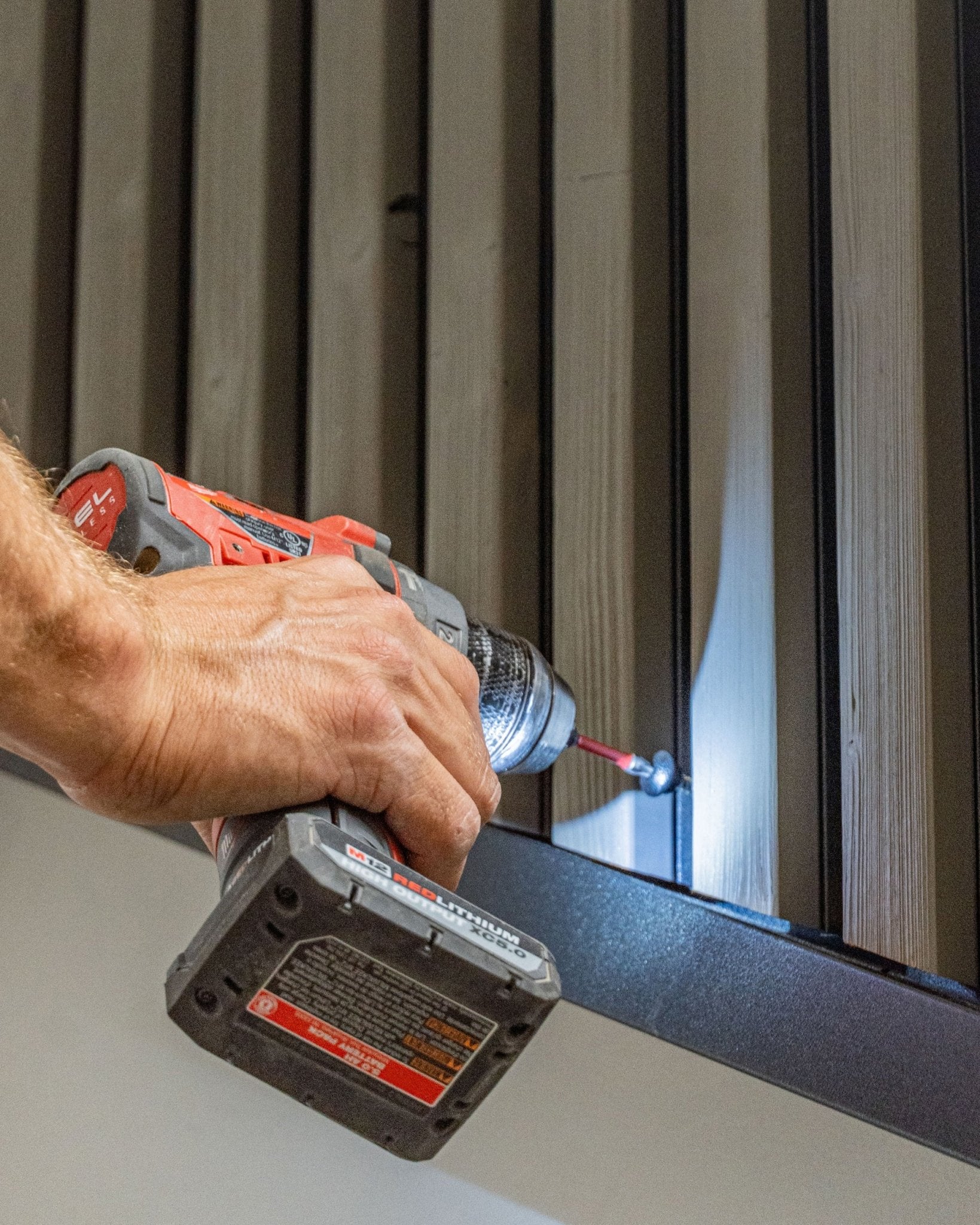 En person fester en black WoodUpp Front Screw (4.8x35, Ø9.5-skive) i et vertical Aluwood fasadepanel med en batteridrill.