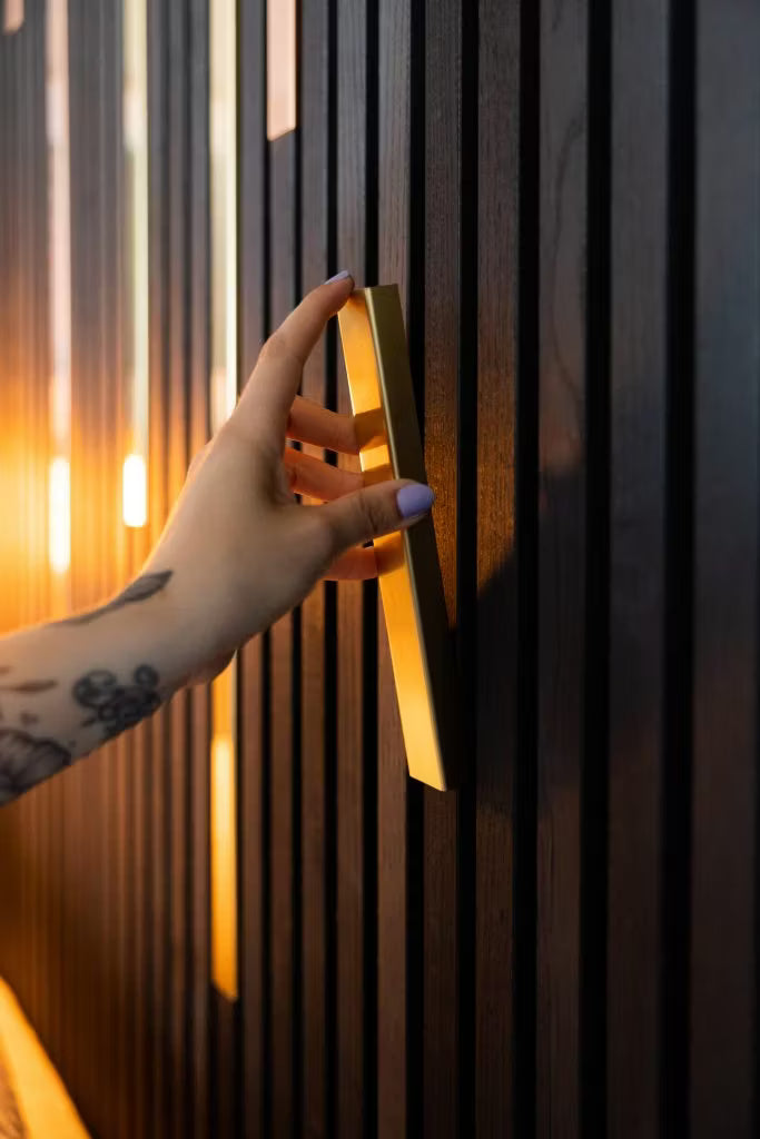 A tattooed hand with light purple nails grips a gold handle on a sleek, dark Akupanel acoustic wood panel wall, highlighted by warm lighting.