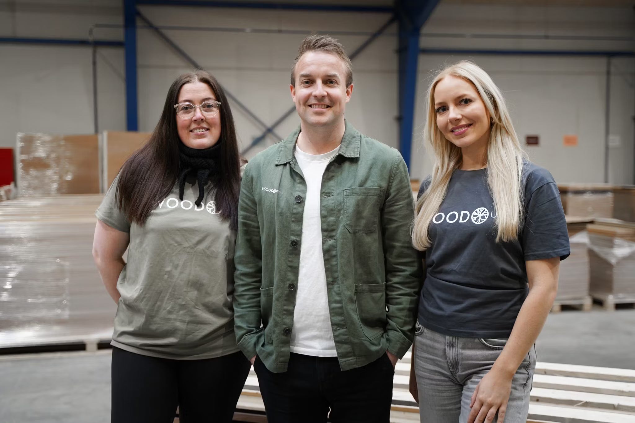 Three people stand and smile in a warehouse, two wearing WOOD branded t-shirts and one in a green jacket, near Nordic-style acoustic panels.