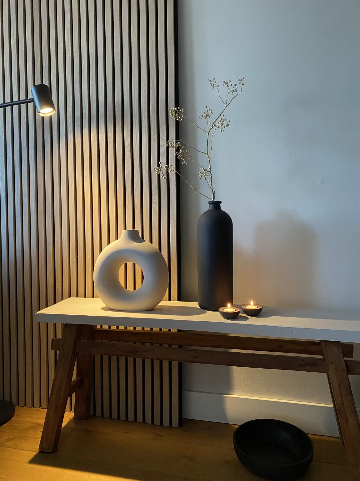 Minimalist decor featuring a white circular vase, black tall vase with dried stems, two tealight candles, and a black bowl set against an Akupanel wood slat acoustic panel backdrop.