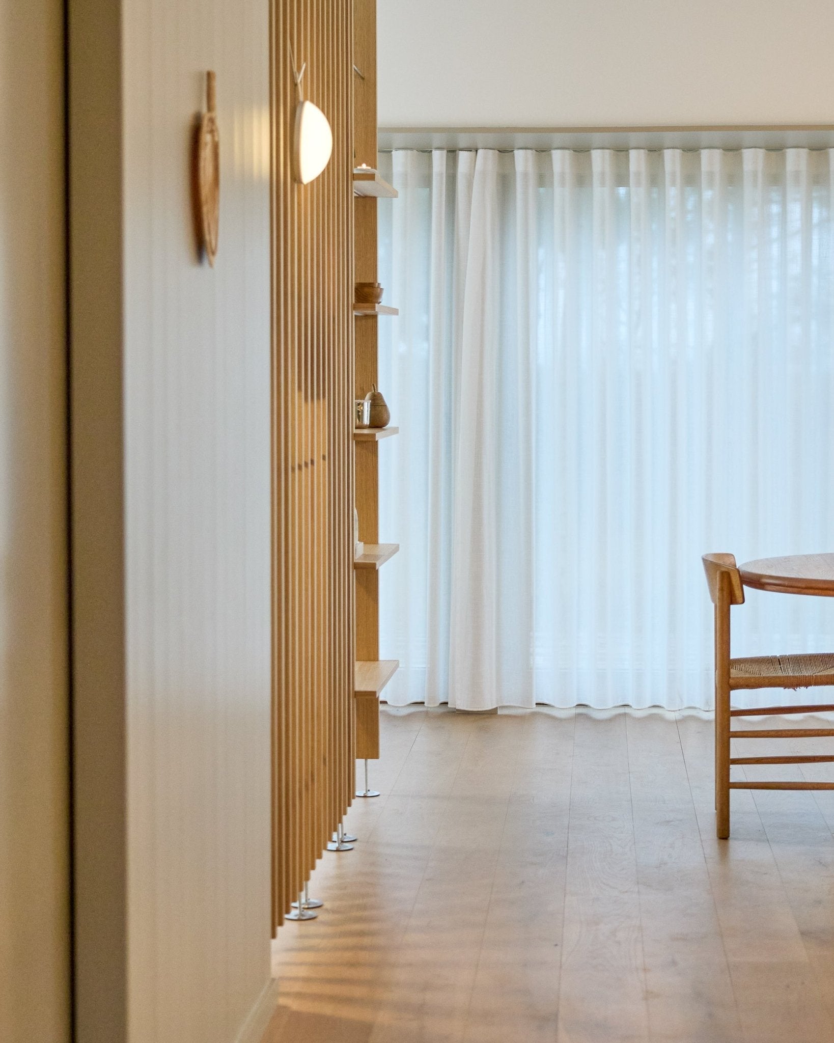 Minimalistisk rom med en gulv-til-tak-romdeler fra WoodUpp i Embrace, tregulv, hyller, stol og lette gardiner foran large vinduer.