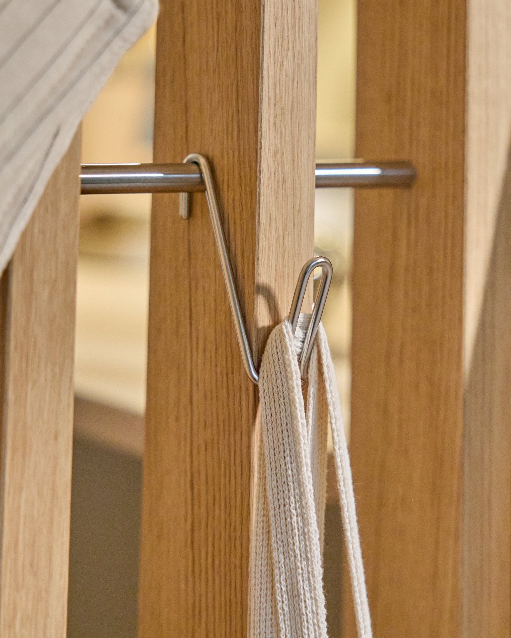 Alva Wire Hook / BM1 (rustfritt Steel, kort) fra WoodUpp er montert på en Akustikkpanel skinne i tre og holder en hvit stropp med metallstang, mot en beige bakgrunn.