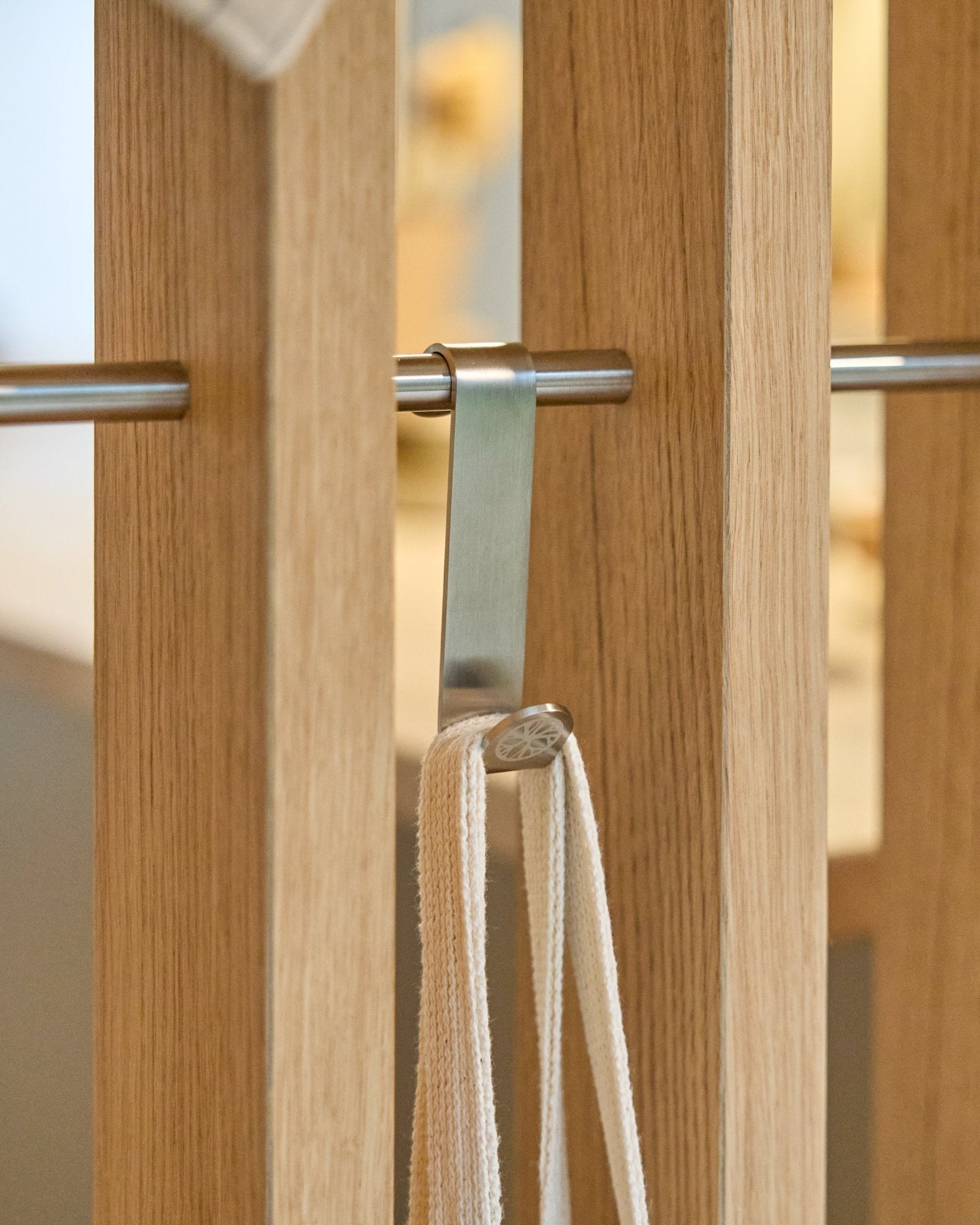 En Alva S-krok fra WoodUpp i rustfritt steel fester en beige stropp til en stang montert mellom vertical akustikkpaneler i tre med spiler. Settet inkluderer 2 kroker.