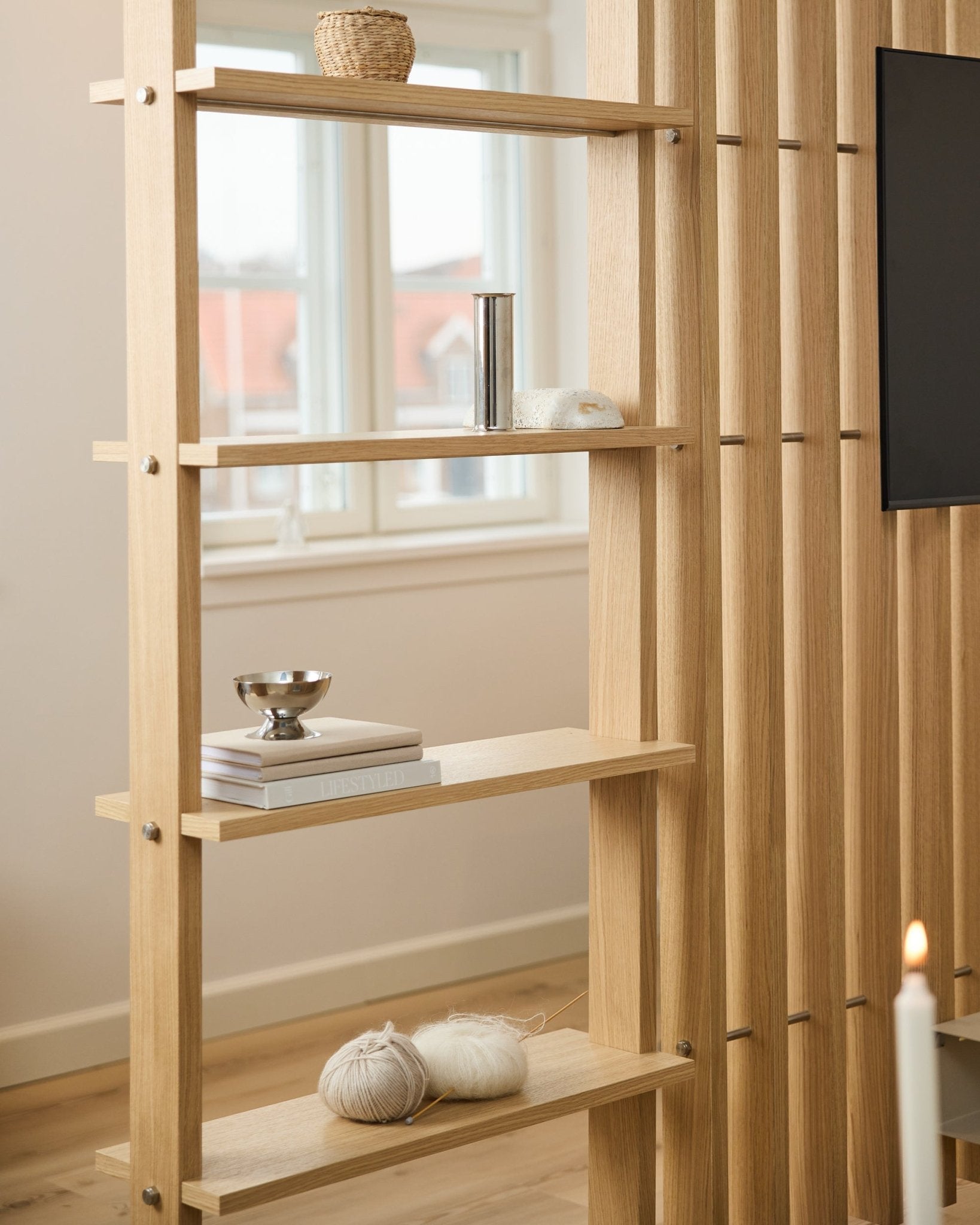 Romdeler i lys eik fra WoodUpp, med vertical spiler og hyller med bøker og garn.