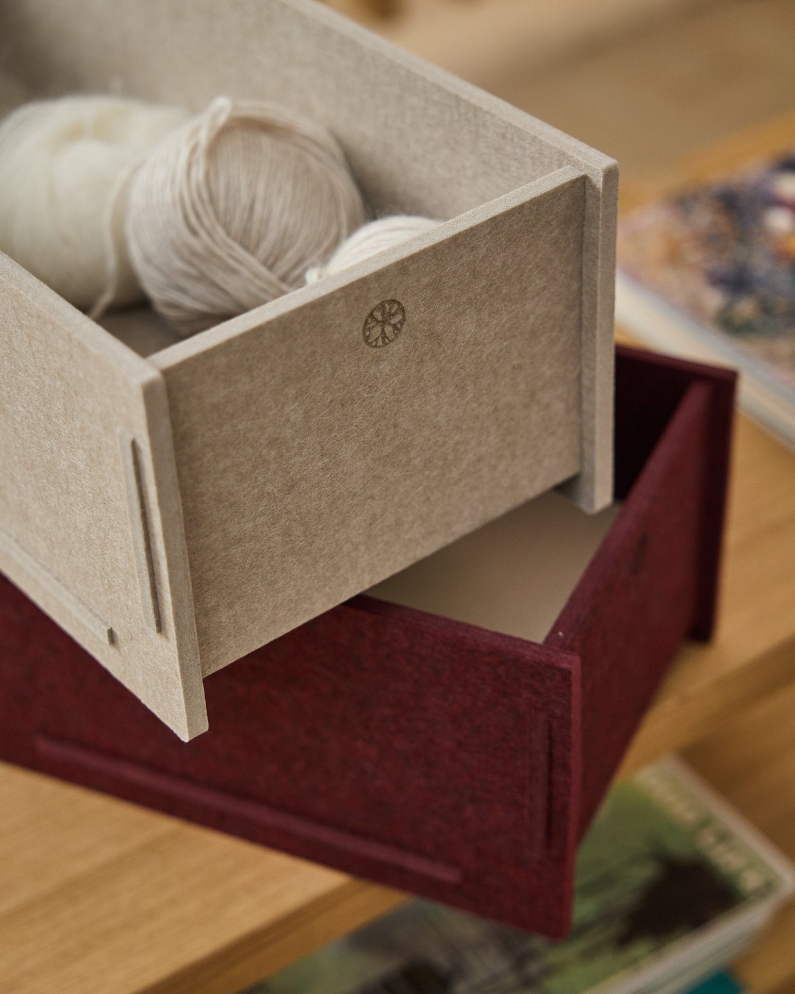 En beige PET-filt oppbevaringsboks fylt med garn står oppå en lukket WoodUpp Vigg Felt Box/BM2 i ox red, plassert på tregulv med bøker i bakgrunnen.