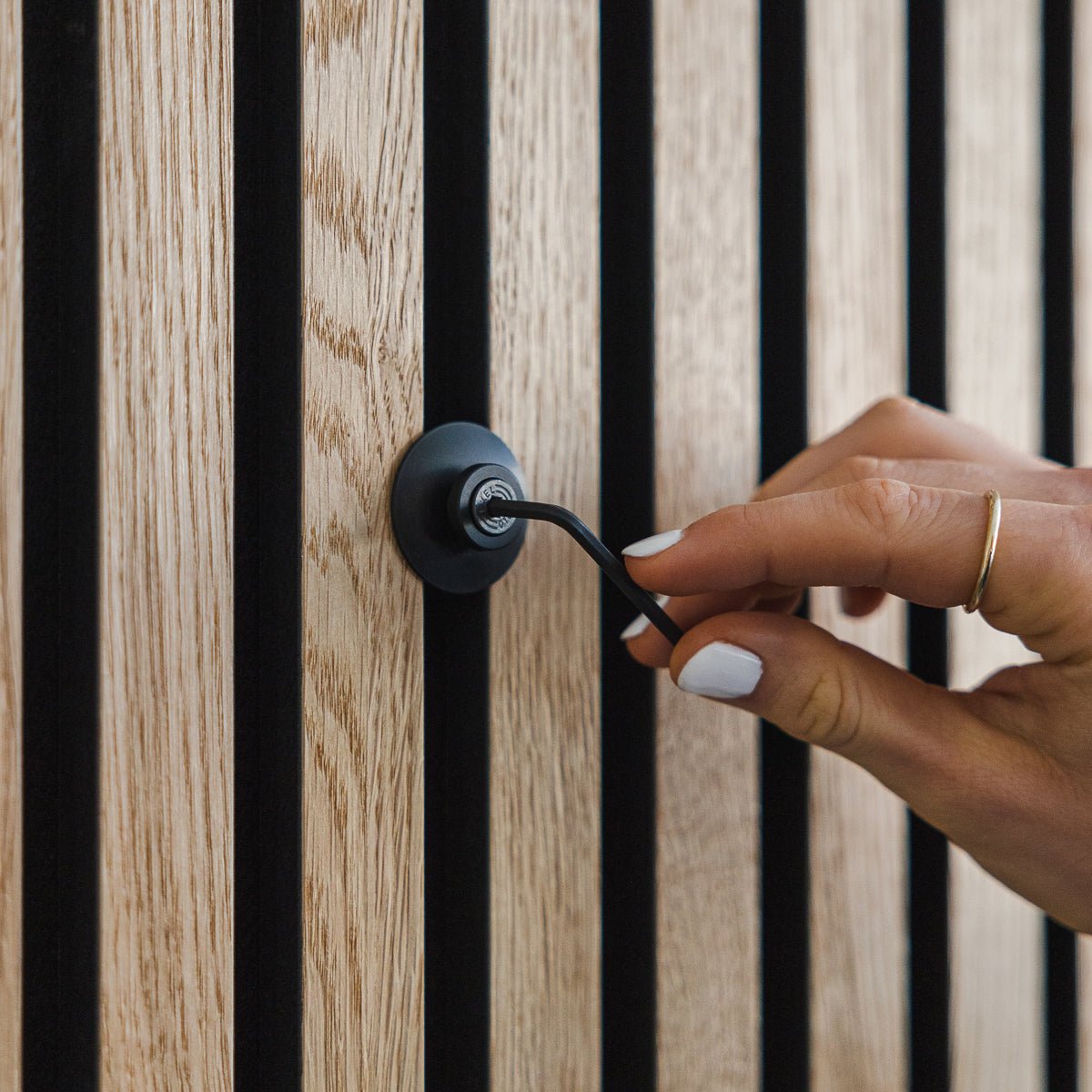 En hånd med hvit neglelakk vrir på en Dupps | Heavy black krok i aluminium fra WoodUpp på et vertical Akupanel akustisk trepanel.