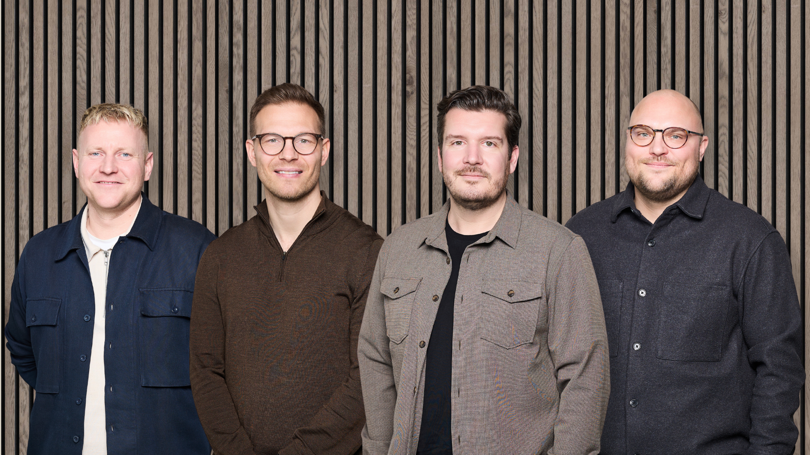 Four men in glasses and casual jackets stand side by side, smiling at the camera in front of a Nordic-style wooden acoustic slatwall panel.